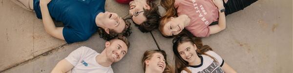 Three women and three men lying down with their heads in a circle smiling.